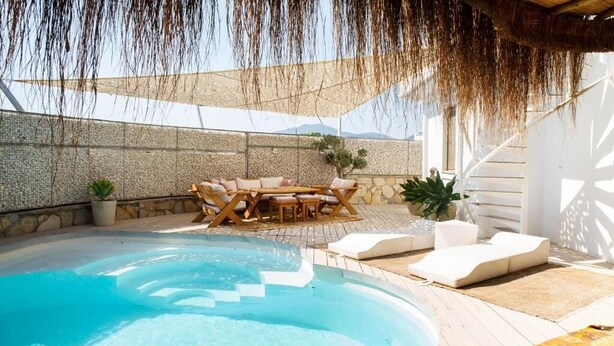 Un área de piscina con terraza de madera, sillas de descanso y una mesa con cojines.