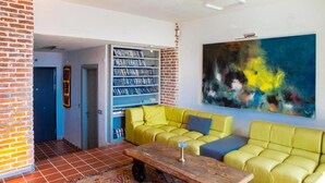 Una sala de estar con una pared de ladrillo, una estantería, un sofá amarillo, un sofá azul y un cuadro abstracto y colorido.