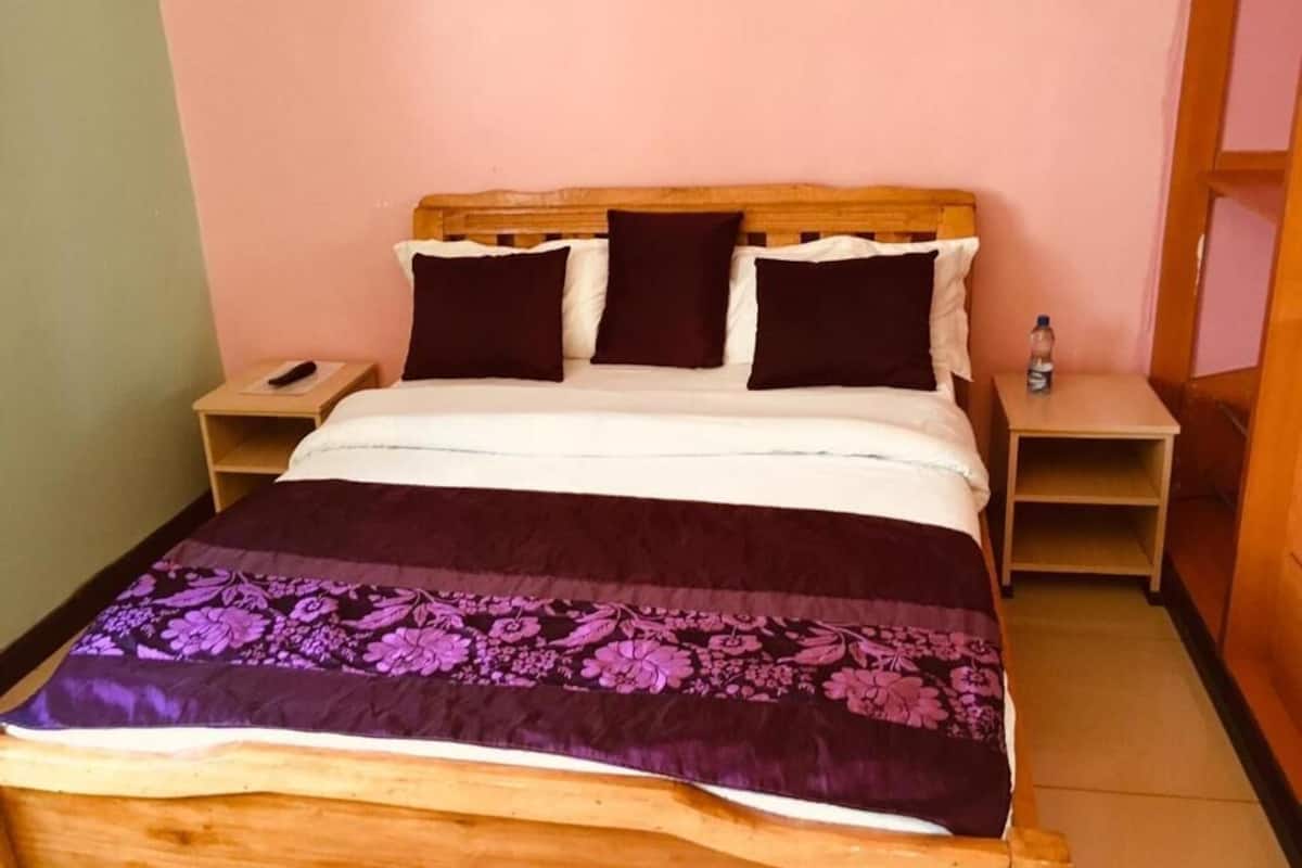 Una cama con cabecera de madera, colcha floral púrpura y dos mesitas de noche, con una botella en una de ellas.