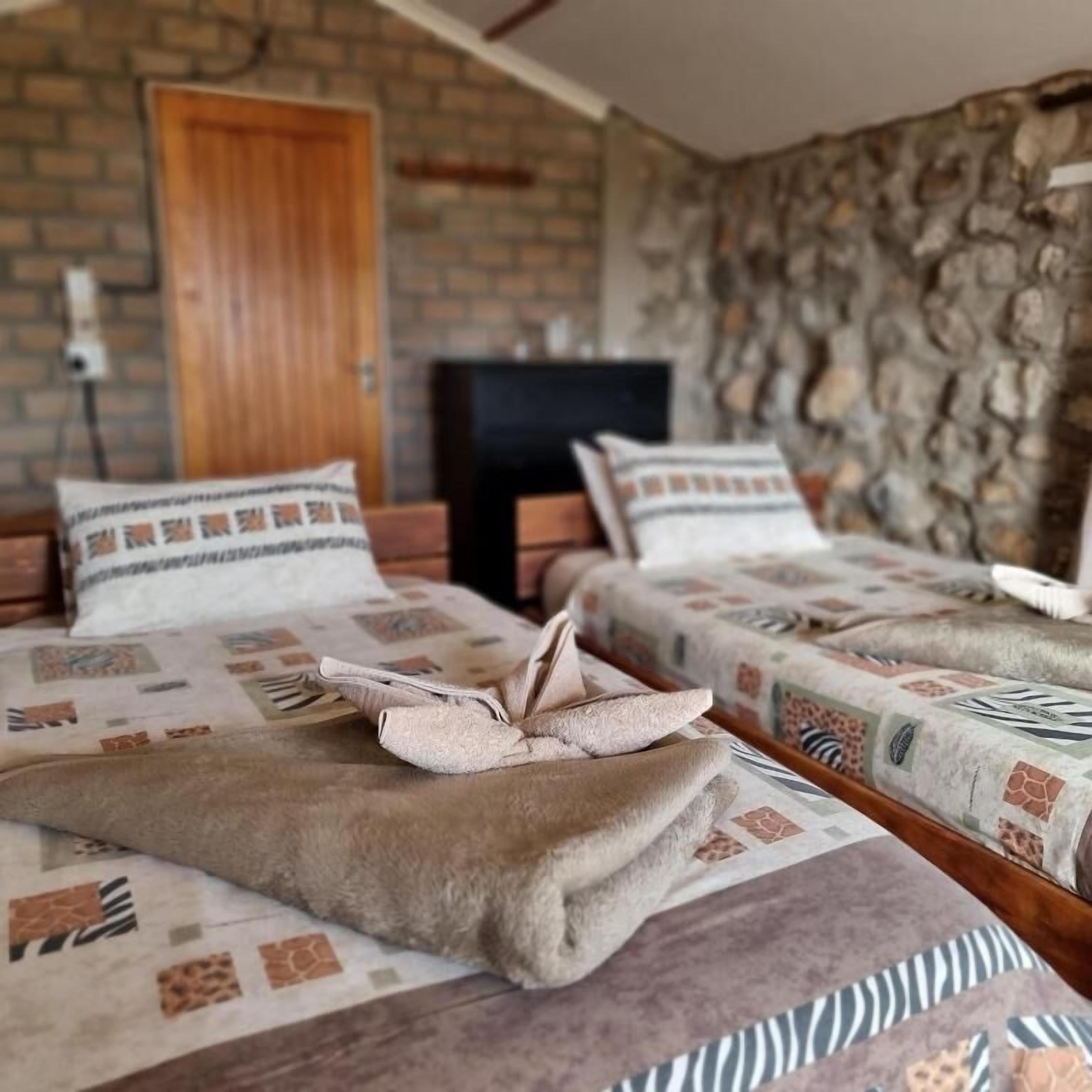 Una habitación con dos camas, cada una con ropa de cama estampada y una manta doblada encima. Se observa una puerta de madera y una pared de piedra al fondo.