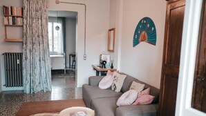 Una sala de estar con un sofá gris, una mesita redonda pequeña y un cojín a cuadros. Se ve una estantería, un radiador y una ventana con cortinas.