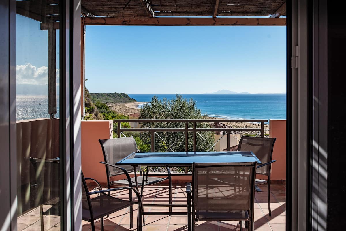 A balcony with a table and chairs, overlooking a beach and the sea.