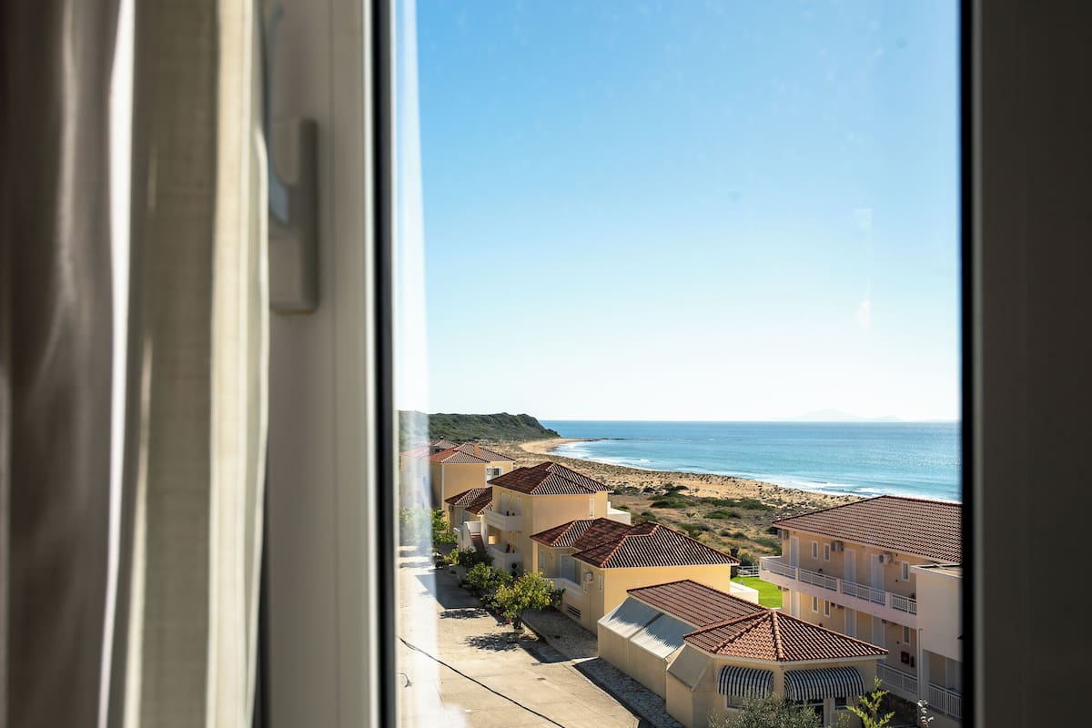A coastal residential area with houses having red-tiled roofs, a beach, and the ocean in the background.