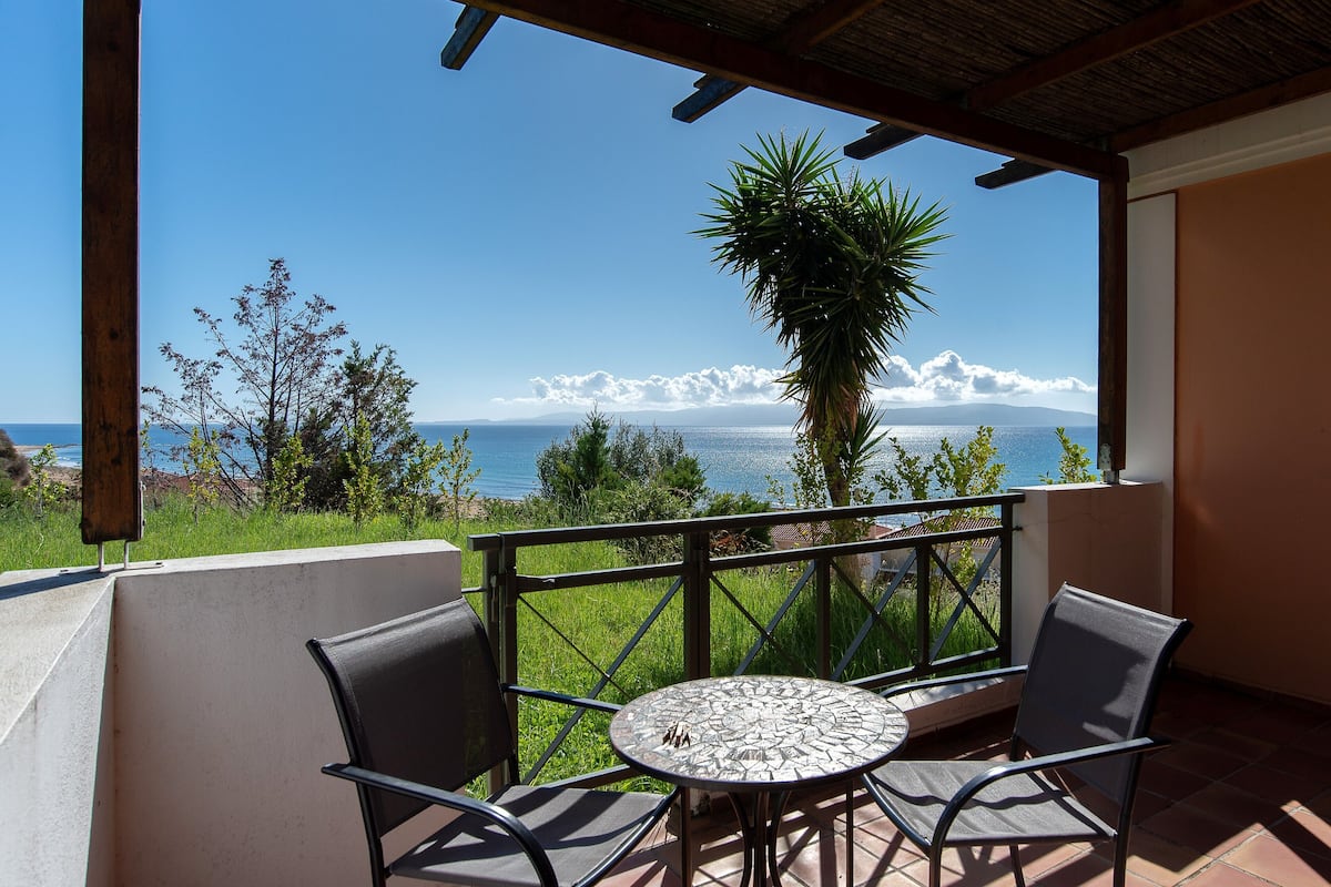 A balcony with a table and chairs, overlooking a scenic view of the sea and mountains.