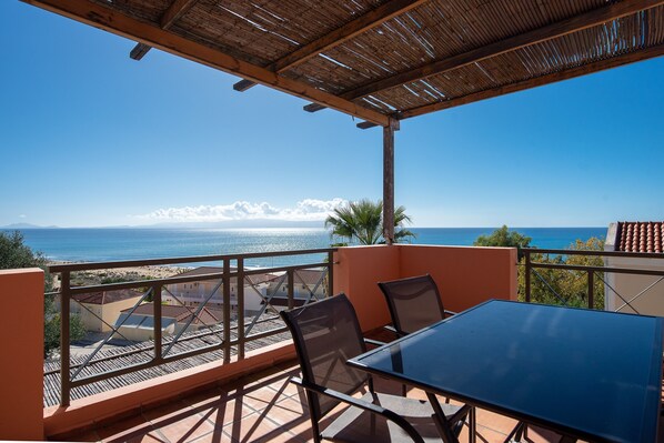 A terrace with a table and chairs, overlooking a coastal view with buildings and the sea. - Mounda Beach Retreat (Skala)