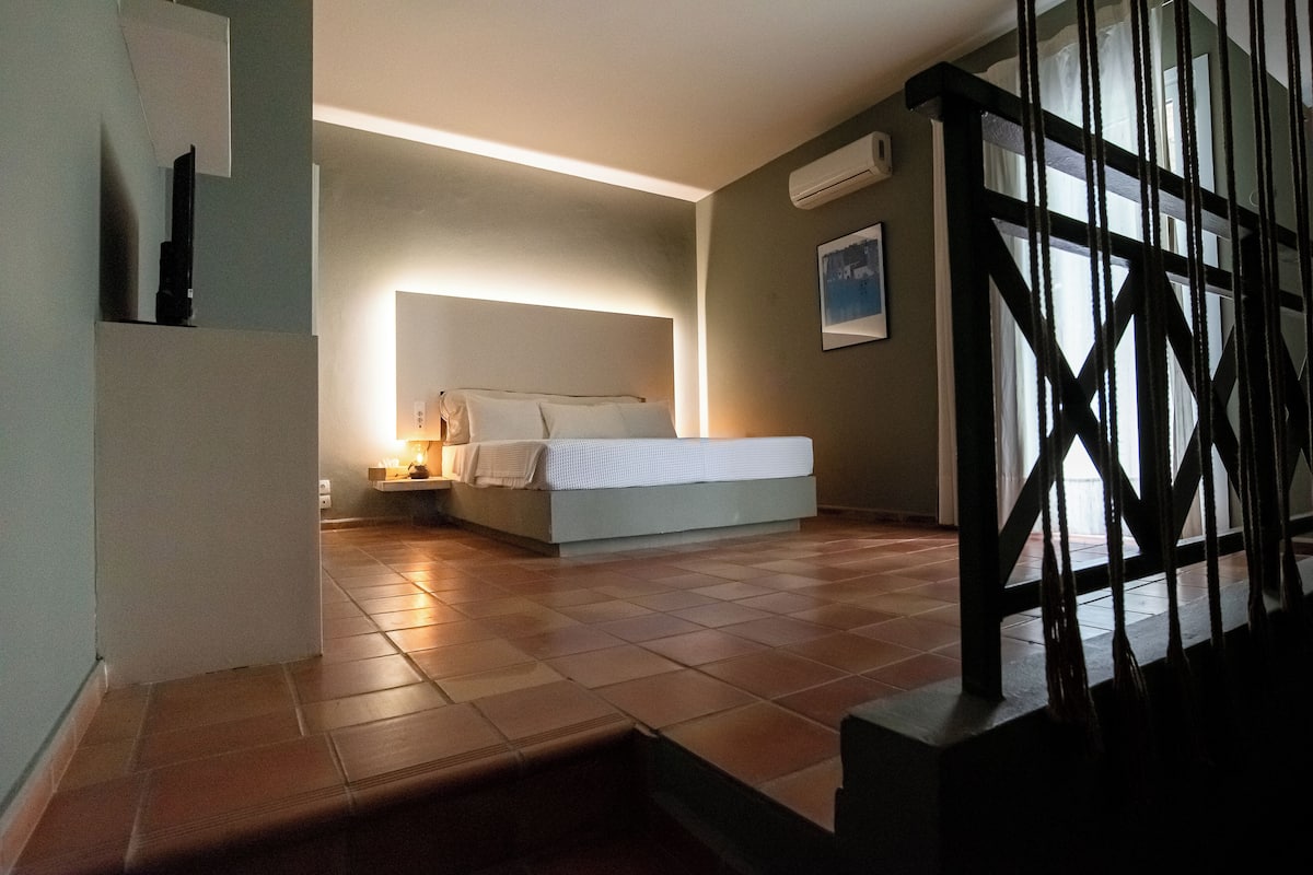A bedroom with a bed, bedside table, and a nightstand. There is a window with curtains, a wall-mounted air conditioner, and a painting on the wall.