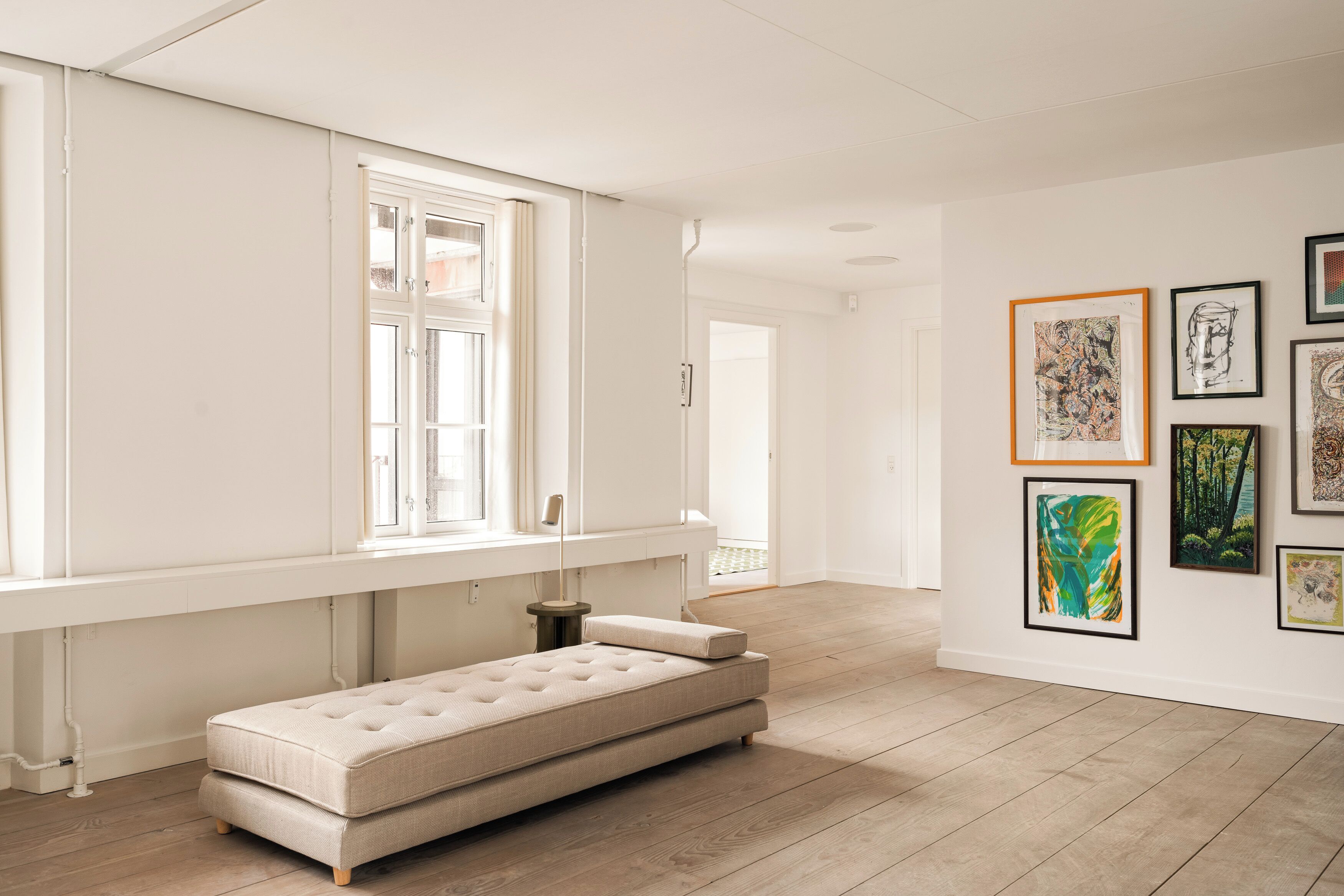 Ein minimalistisch eingerichteter Raum mit einem großen Fenster, einem Sofa und mehreren gerahmten Kunstwerken an der Wand.