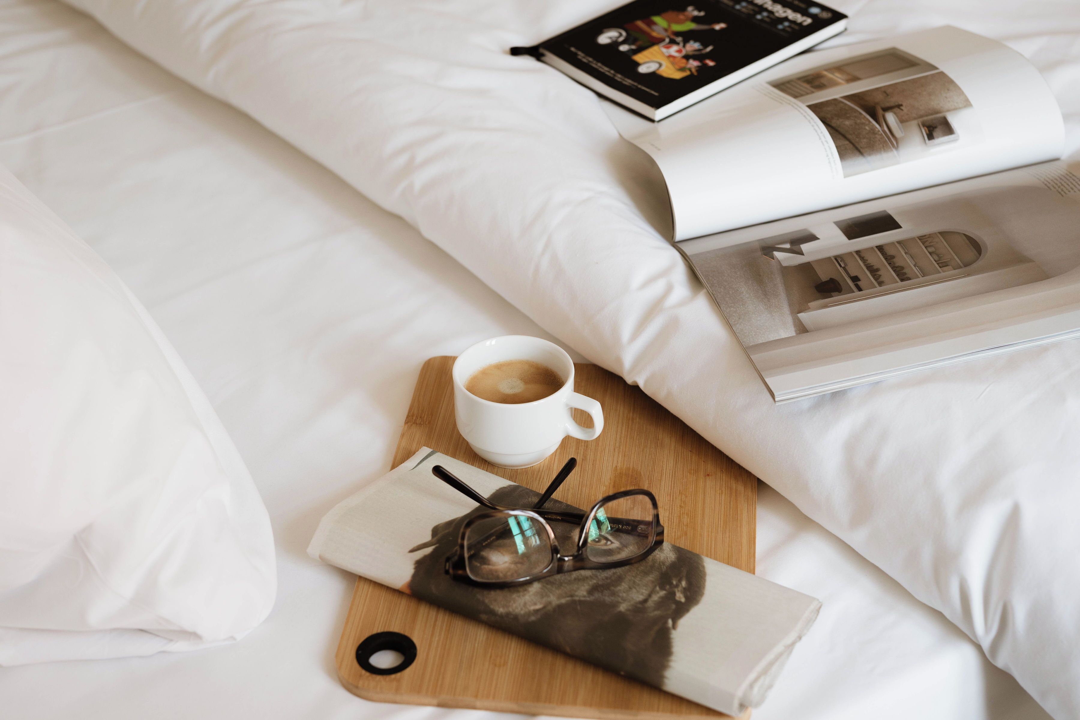 Uma cama bem arrumada com lençóis brancos, uma bandeja de madeira com uma xícara de café, copos e uma revista.