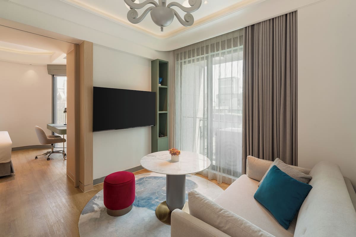 A modern hotel room with a flat-screen TV, a round dining table, a sofa with cushions, a red ottoman, and a chandelier.