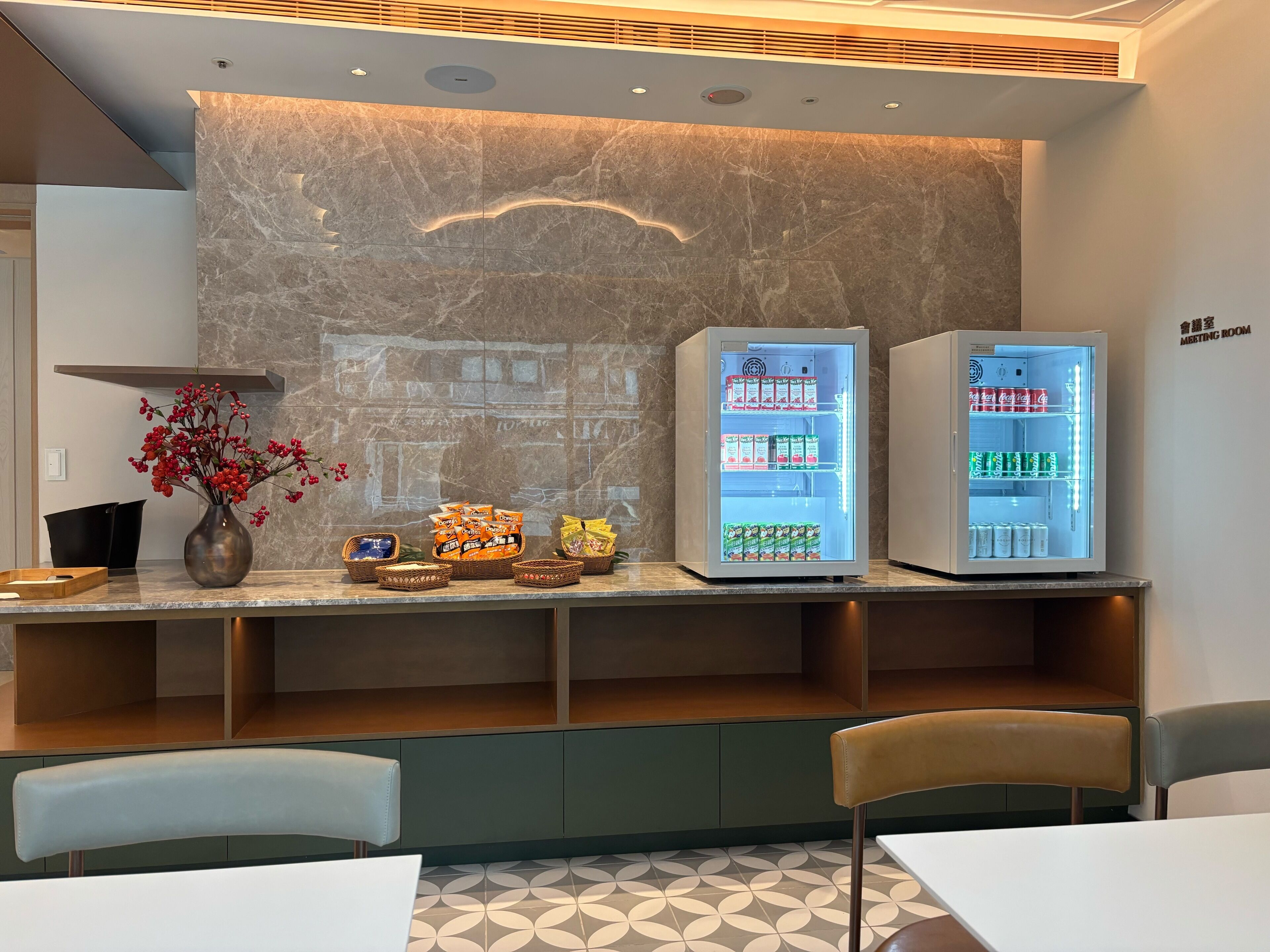 A hotel reception area with marble walls, a reception desk, two illuminated beverage dispensers, and a seating area with chairs and tables.