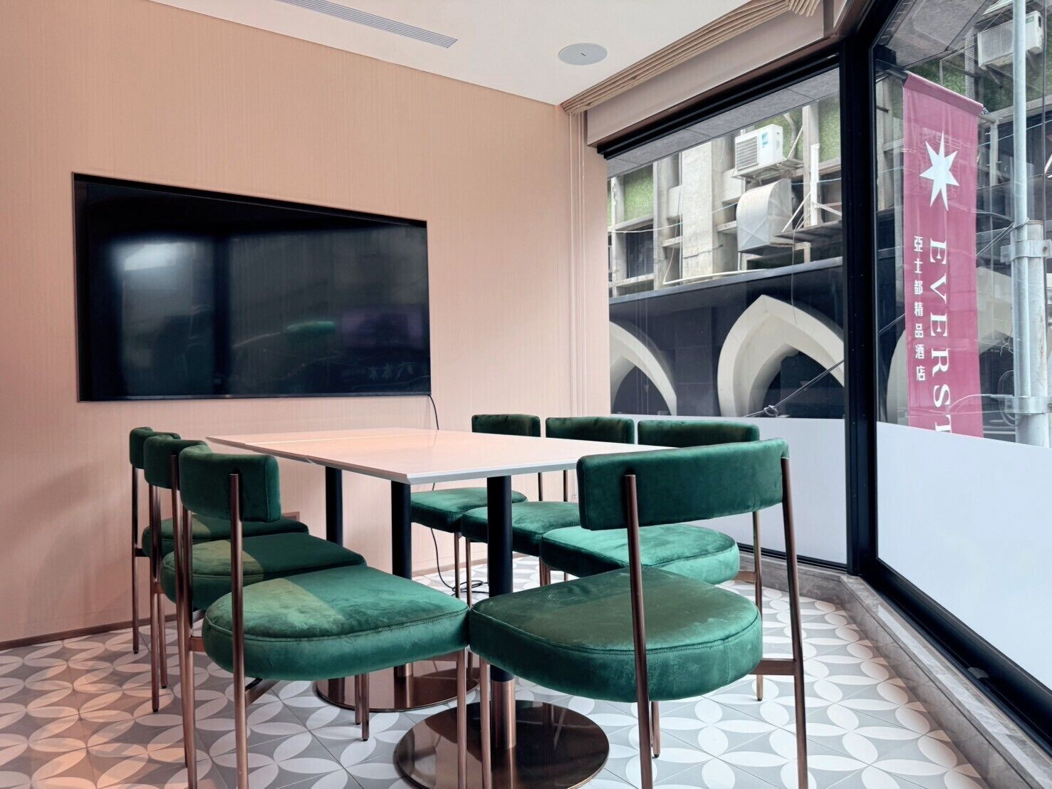 A conference room with a large table and green upholstered chairs, a large screen on the wall, and a view of the outside through a large window.