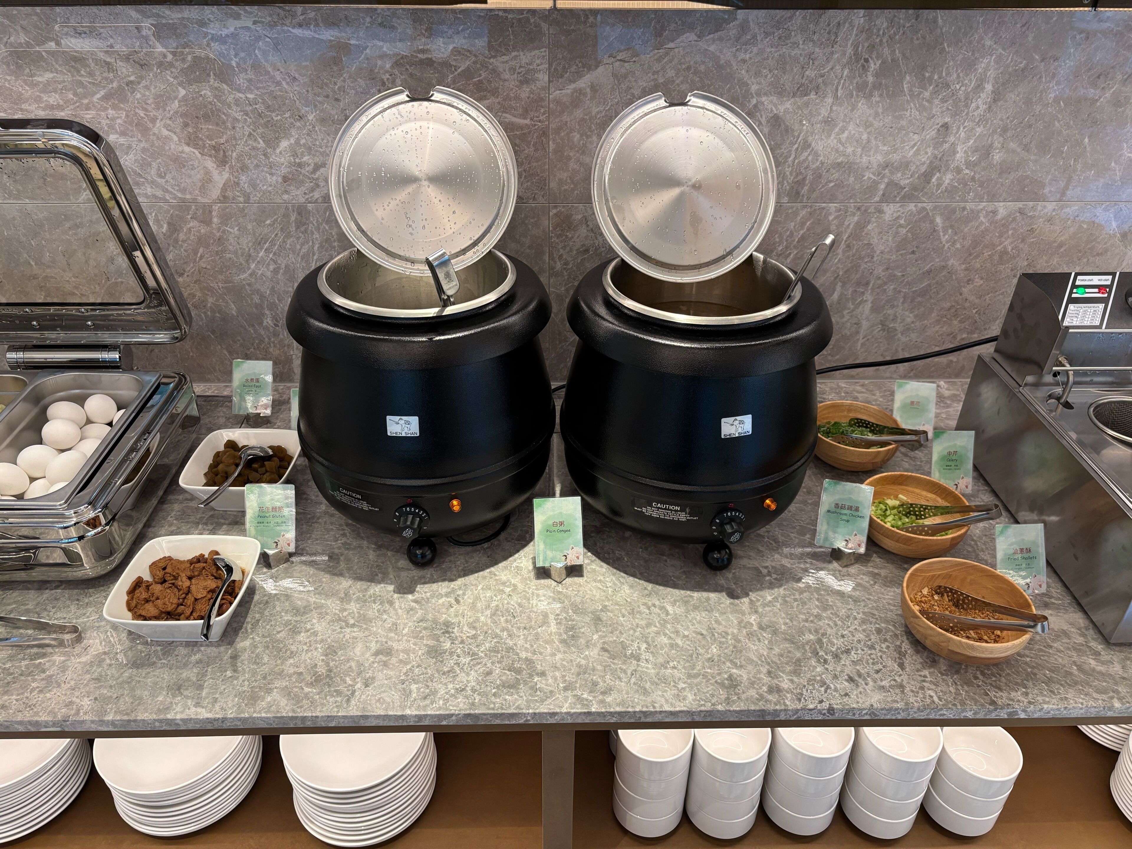 A buffet setup with two large black appliances, a tray of eggs, bowls of food, and stacked white plates.
