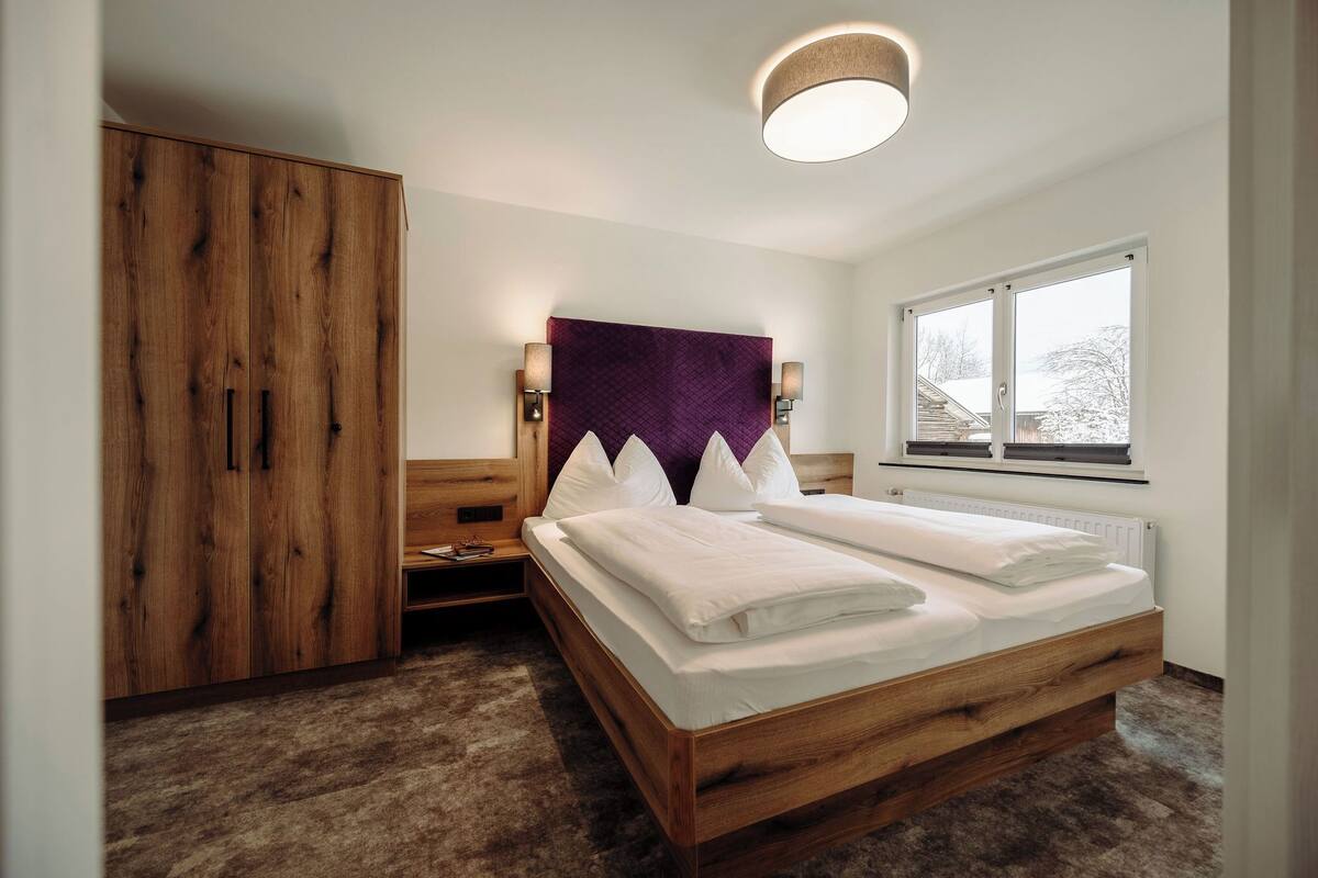 A bedroom with a wooden bed, a wardrobe, a nightstand, a purple headboard, and a window with a view of trees.