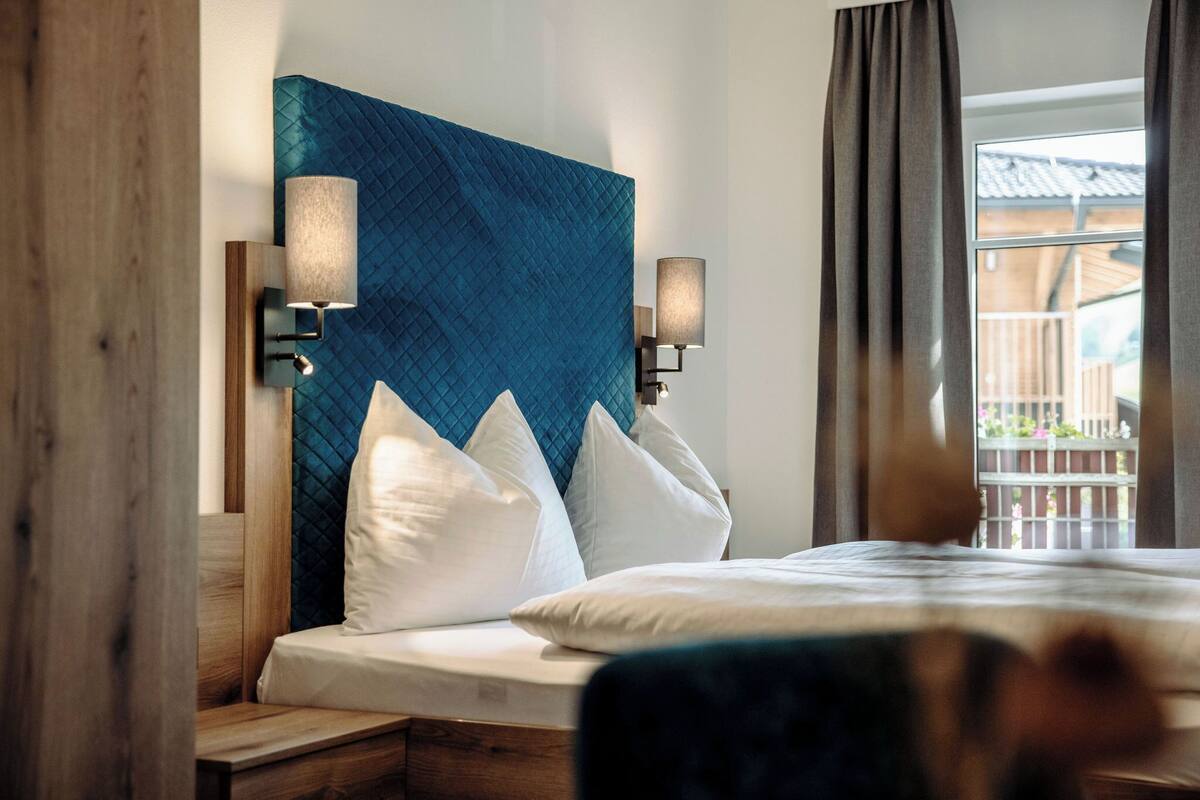 A hotel room with a large bed, two wall-mounted lamps, and a view of the outside balcony.