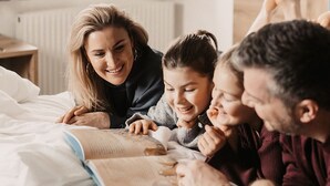 Une famille de quatre personnes allongée sur un lit, avec un livre, dans une pièce dotée d’une fenêtre et d’une lampe.
