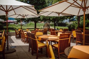 An outdoor dining area with wooden tables and chairs, large umbrellas, and a view of a garden and buildings in the background. - Lerchs Landhotel (Sankt Johann im Pongau)