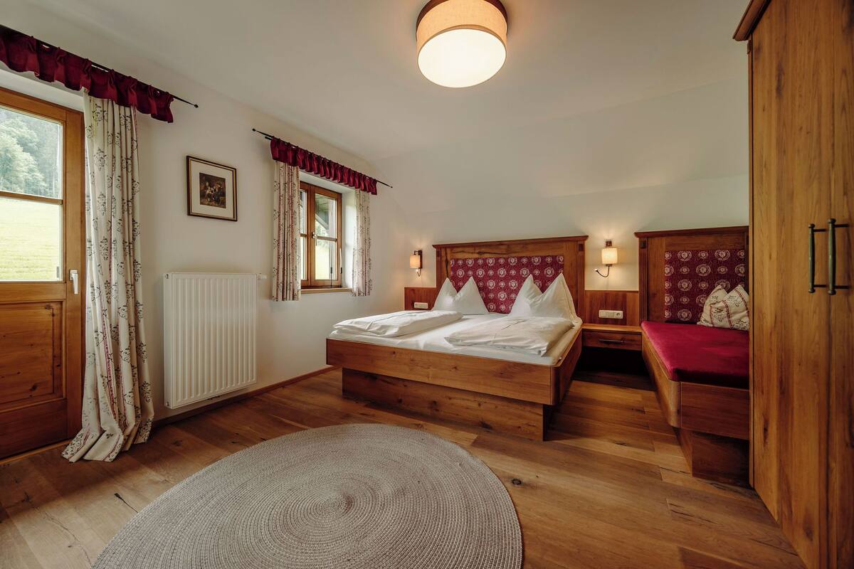 A bedroom with a wooden bed, a bedside table, a red bench, a radiator, and a window with curtains.