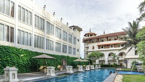 A swimming pool with lounge chairs and umbrellas, surrounded by a lush green hedge and a multi-story building with balconies.