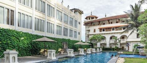 A swimming pool with lounge chairs and umbrellas, surrounded by a lush green hedge and a multi-story building with balconies.