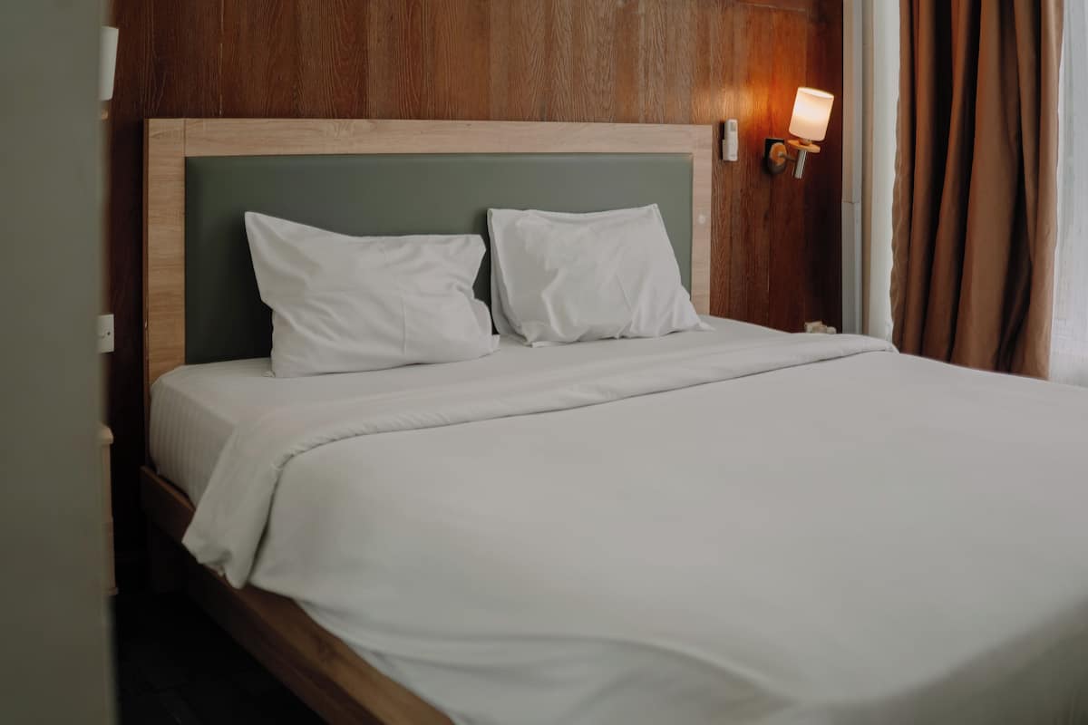 A neatly made bed with white linens and pillows, a wooden headboard, and a wall-mounted light fixture.