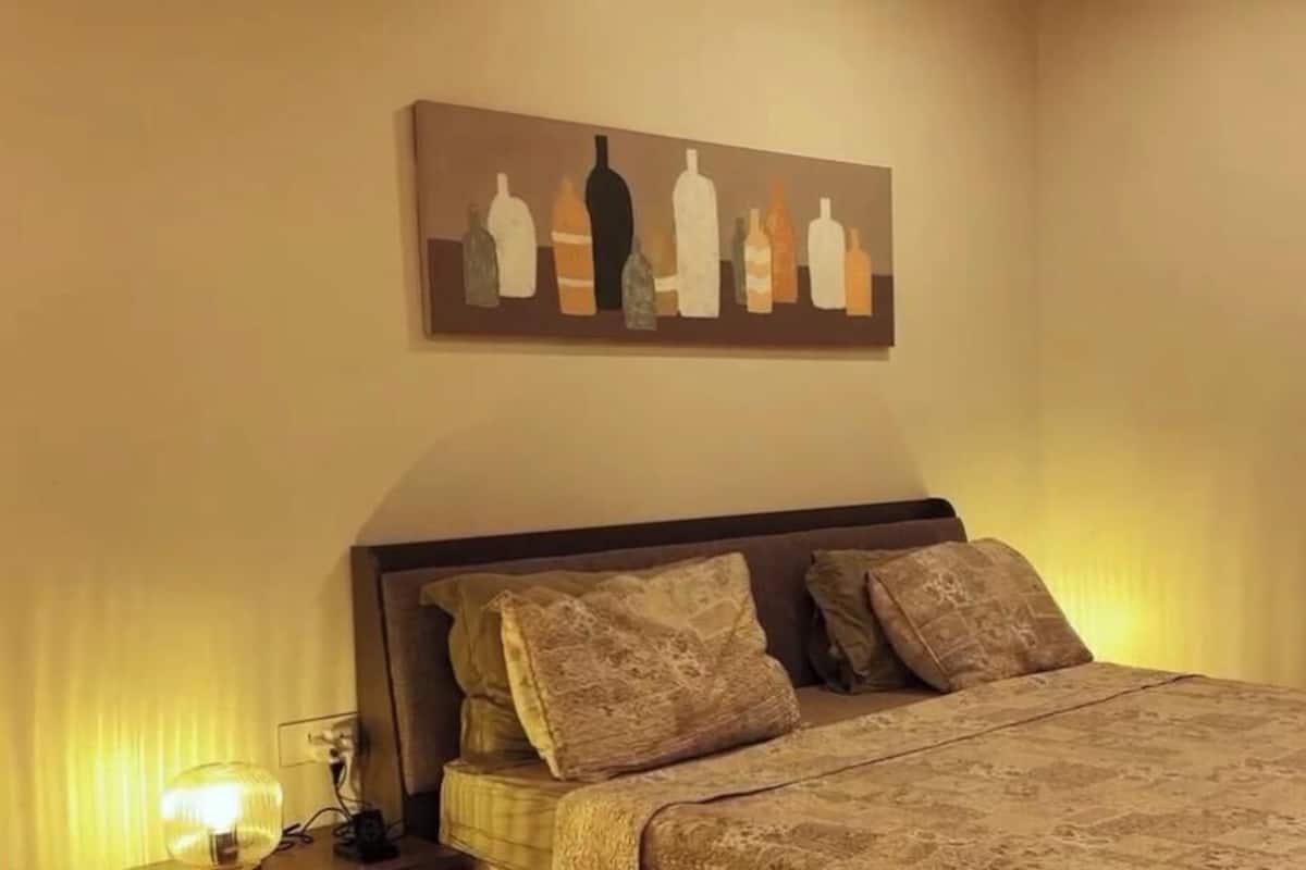 Un dormitorio con cama, almohadas, una mesita de noche, lámpara y un cuadro en la pared.