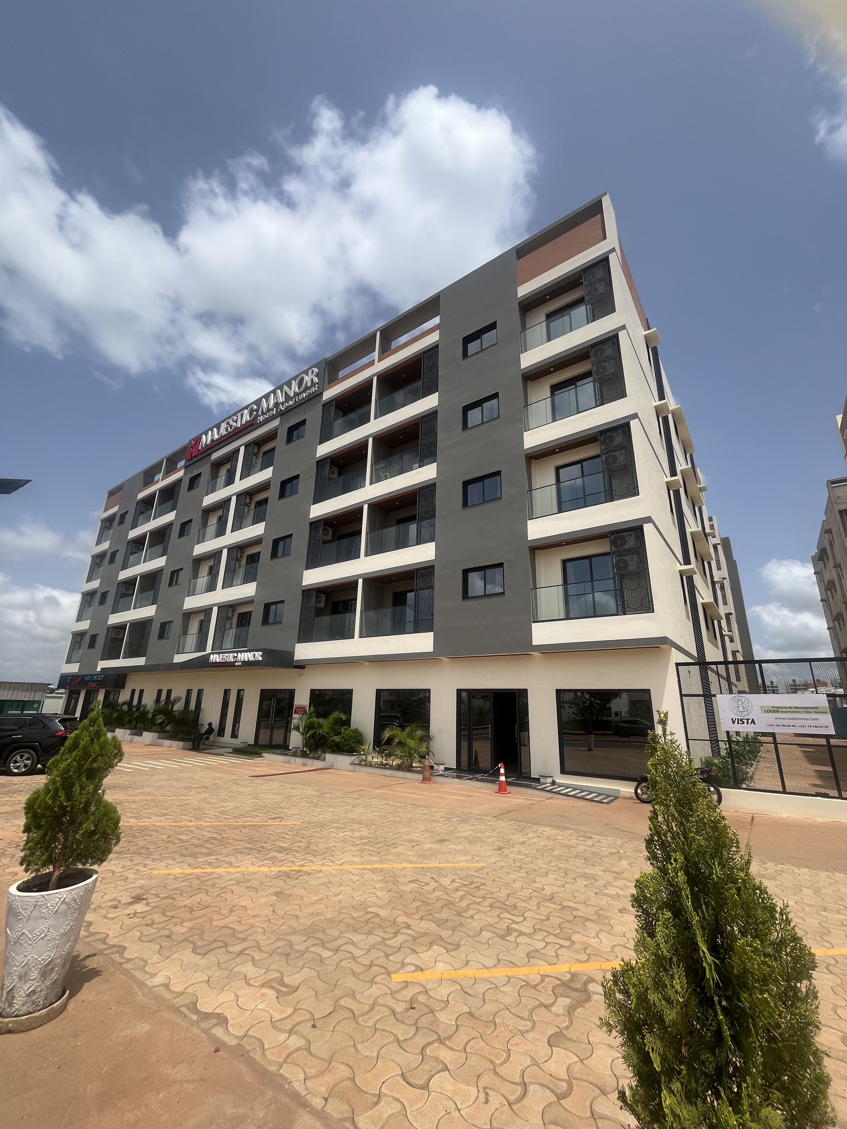 Um edifício de vários andares com varandas, área de entrada pavimentada e uma placa com o nome 'Music Manor'.