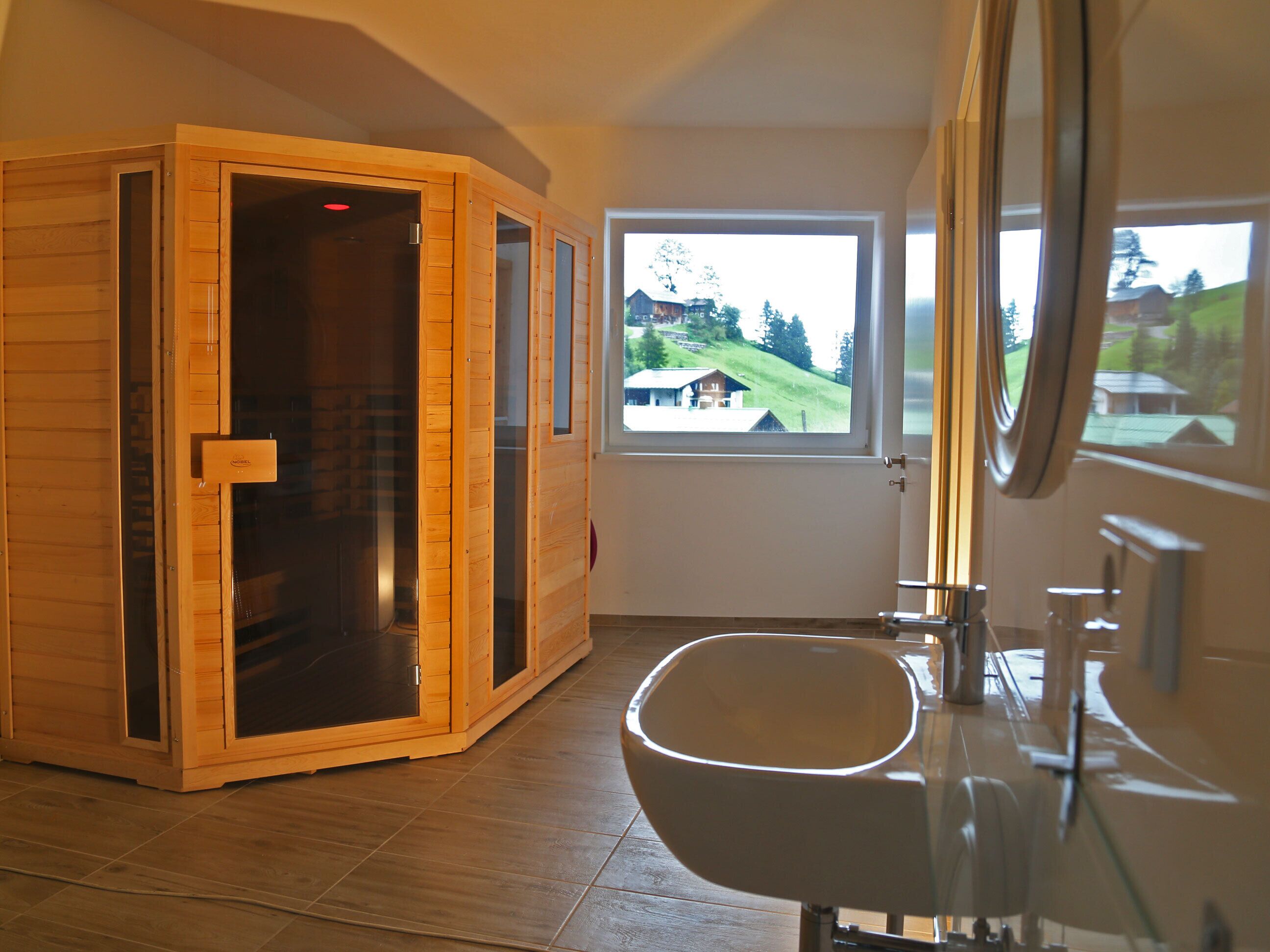 A sauna room with a wooden door, a large window with a view of a green landscape, and a modern bathroom with a sink and mirror.