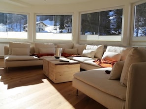 A modern living room with a beige sofa set, a wooden coffee table, and large windows with a view of a snowy landscape. - Berghaus Anna Lisa (Mittelberg)