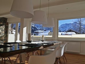 A modern dining area with a glass table, white chairs, and pendant lights. A stone wall and a window with a mountain view are visible. - Berghaus Anna Lisa (Mittelberg)
