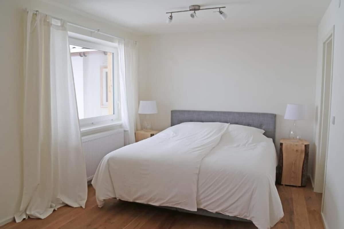 Un dormitorio con una cama, dos mesitas de noche, una ventana con cortinas y una lámpara en el techo.