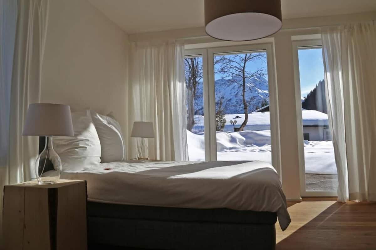 Un dormitorio con cama, dos lámparas de noche, una mesita de noche de madera y vistas a un paisaje nevado a través de amplios ventanales.