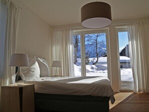 Ein Schlafzimmer mit einem Bett, zwei Nachttischlampen, einem Holznachttisch und Blick auf eine verschneite Landschaft durch große Fenster.