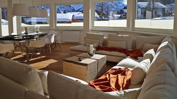 A modern living room with a sofa, coffee table, and dining area. Large windows offer views of a snowy landscape. - Berghaus Anna Lisa (Mittelberg)