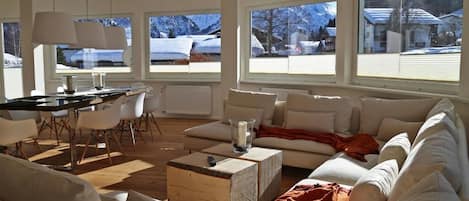 Una sala de estar moderna con sofá, mesa de centro y comedor. Amplias ventanas ofrecen vistas a un paisaje nevado.