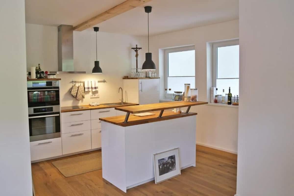 Una cocina moderna con armarios blancos, una isla de madera, iluminación colgante y vistas al exterior a través de una ventana.