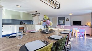 Une cuisine moderne avec des meubles blancs, un coin repas avec une table en bois dressée pour quatre personnes, et un salon avec un canapé et une télévision.