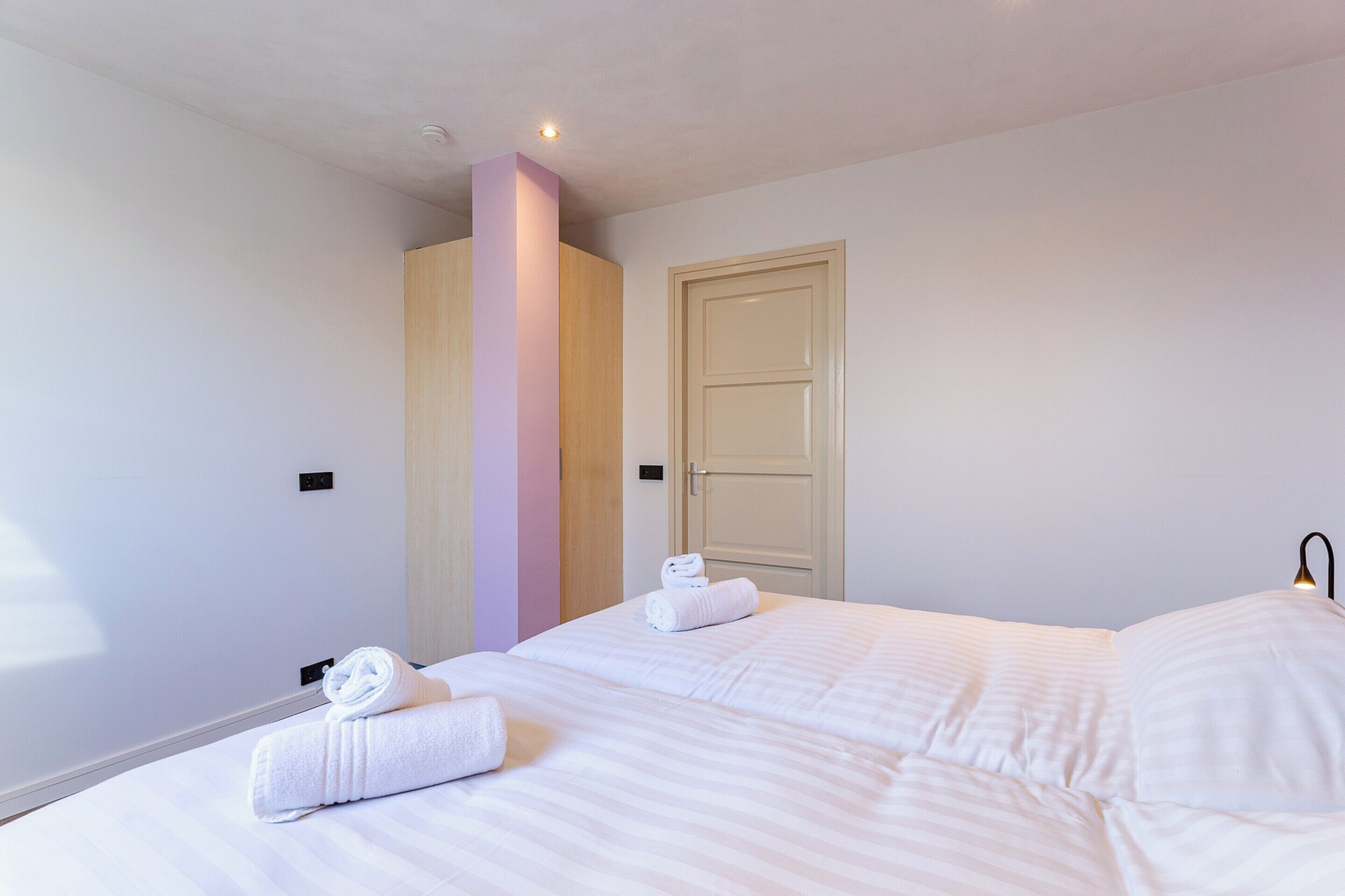 A hotel room with a bed, two rolled towels, a door, and a light fixture.