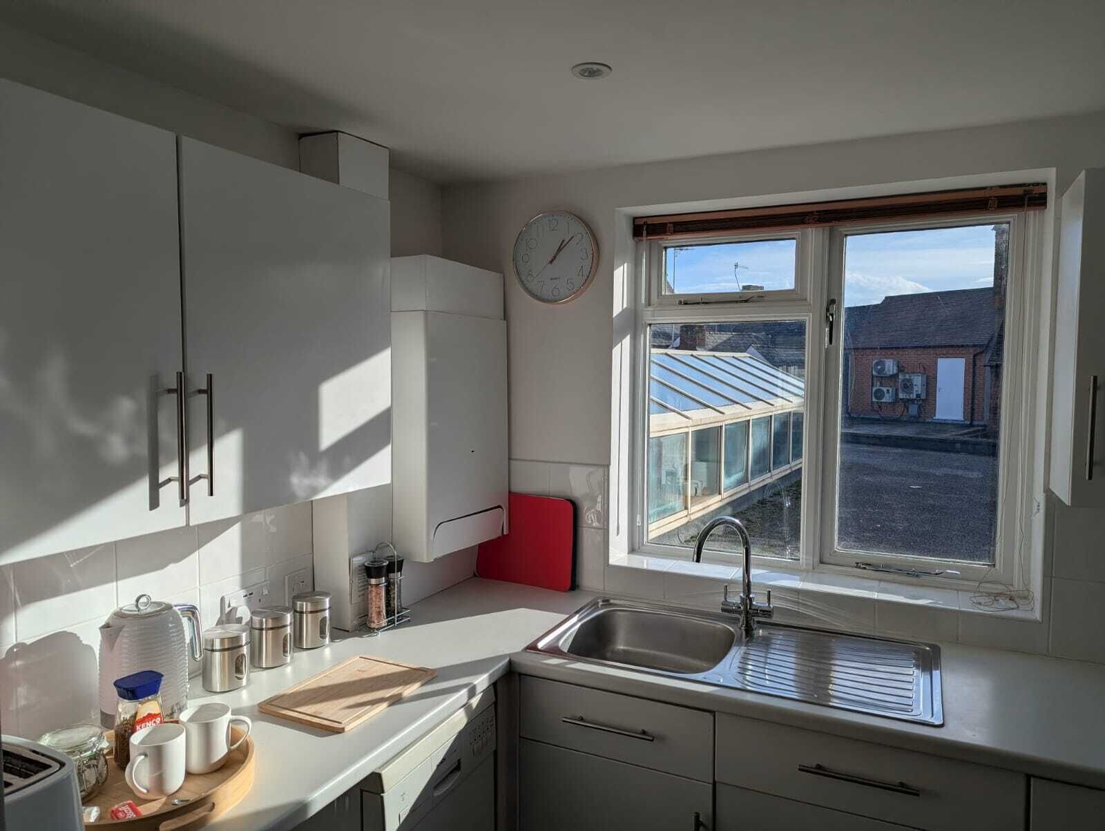 Eine Küche mit weißen Schränken, einer roten Arbeitsplatte, einem Edelstahl-Spülbecken und einem Fenster mit Blick auf ein Gewächshaus und Gebäude.