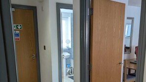 Un couloir avec des portes en bois, un paillasson noir et une salle de bain visible, équipée d’un lavabo et dont les murs sont recouverts de carreaux.