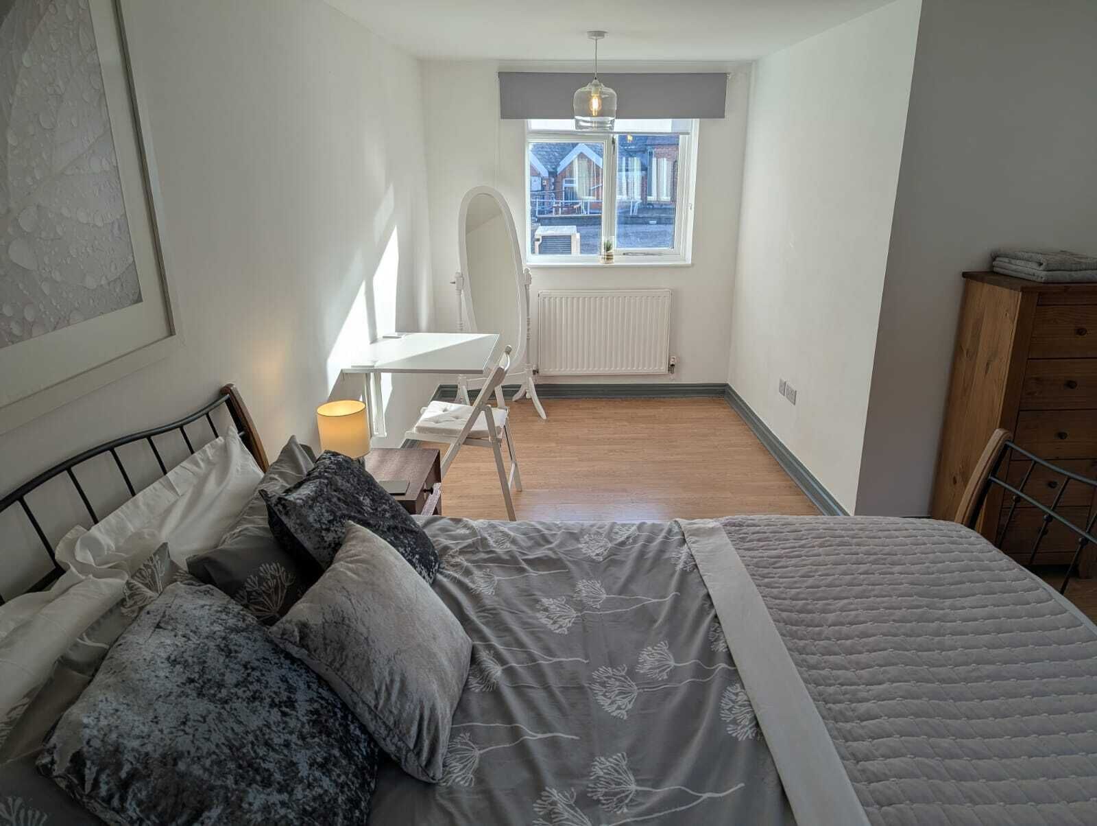 A bedroom with a bed, a desk, a chair, a radiator, and a window with a view of buildings outside.