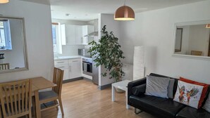 Un appartement moderne comprenant un coin repas, un salon avec un canapé en cuir noir et une cuisine dotée de meubles blancs et d’une grande plante.