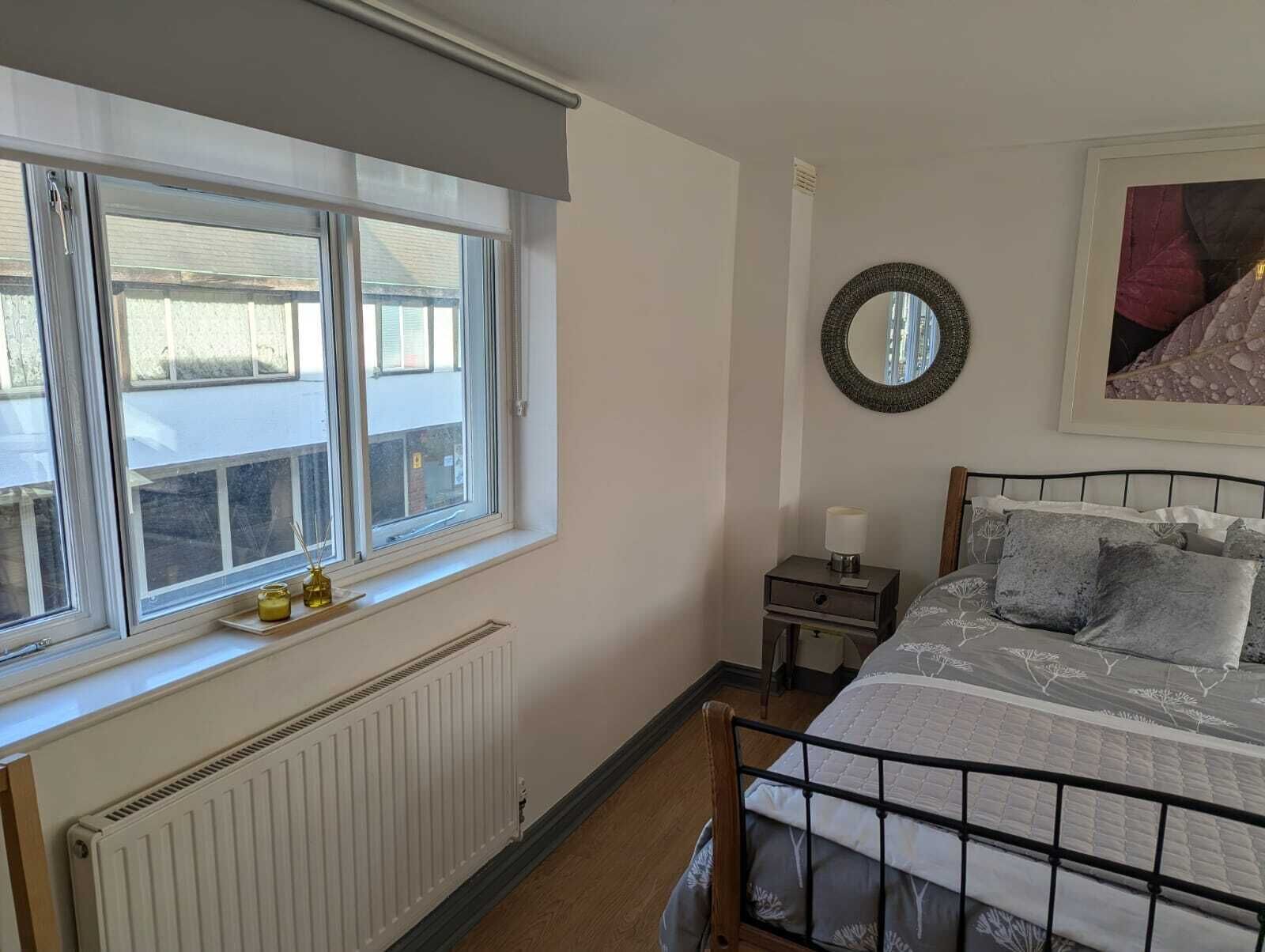 A bedroom with a bed, a bedside table with a lamp, a window with a view of buildings, a radiator, and a framed picture on the wall.