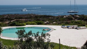 En swimmingpool med en person som simmar, omgiven av solstolar och med utsikt över havet och båtar.