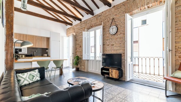 A modern living room with a black leather sofa, a coffee table, and a TV. There is a kitchen in the background with wooden cabinets and a dining area with a green chair. - Elegant and bright apartment in the center (Granada)