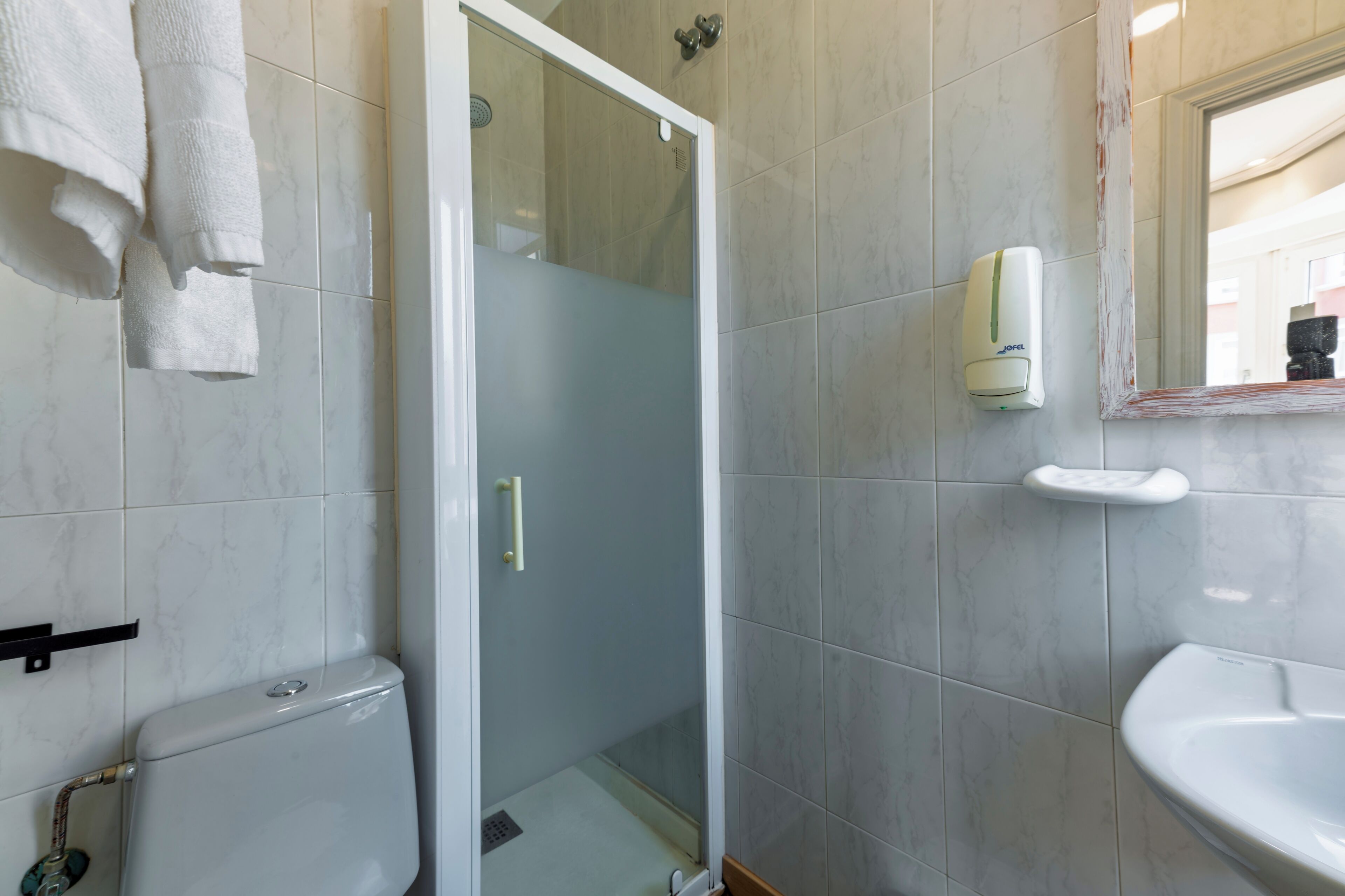 Casa de banho com sanita, chuveiro com porta de vidro, dispensador de sabonete líquido e lavatório.