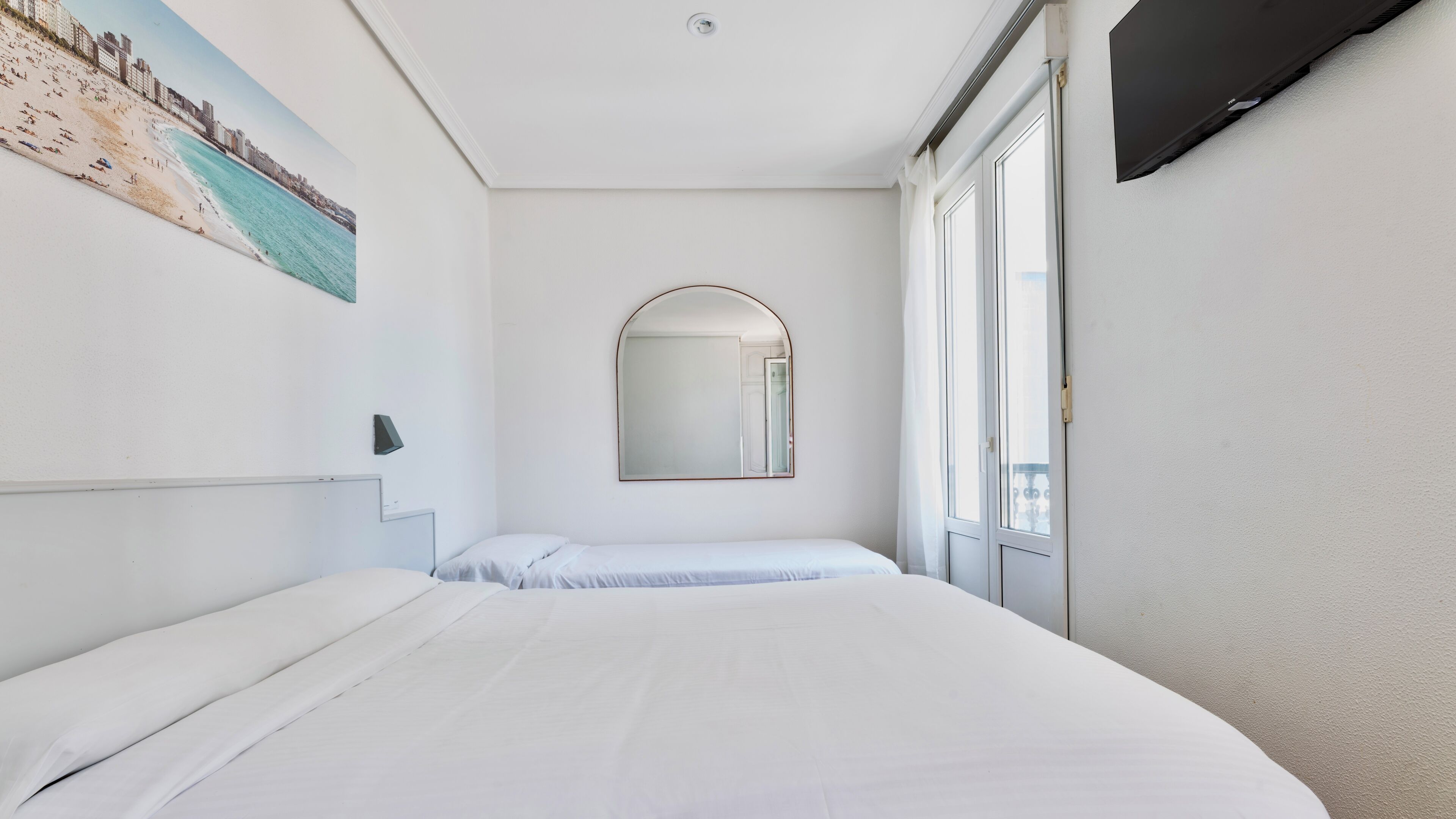 A hotel room with a large bed, a wall-mounted TV, a painting of a beach, and a window with curtains.