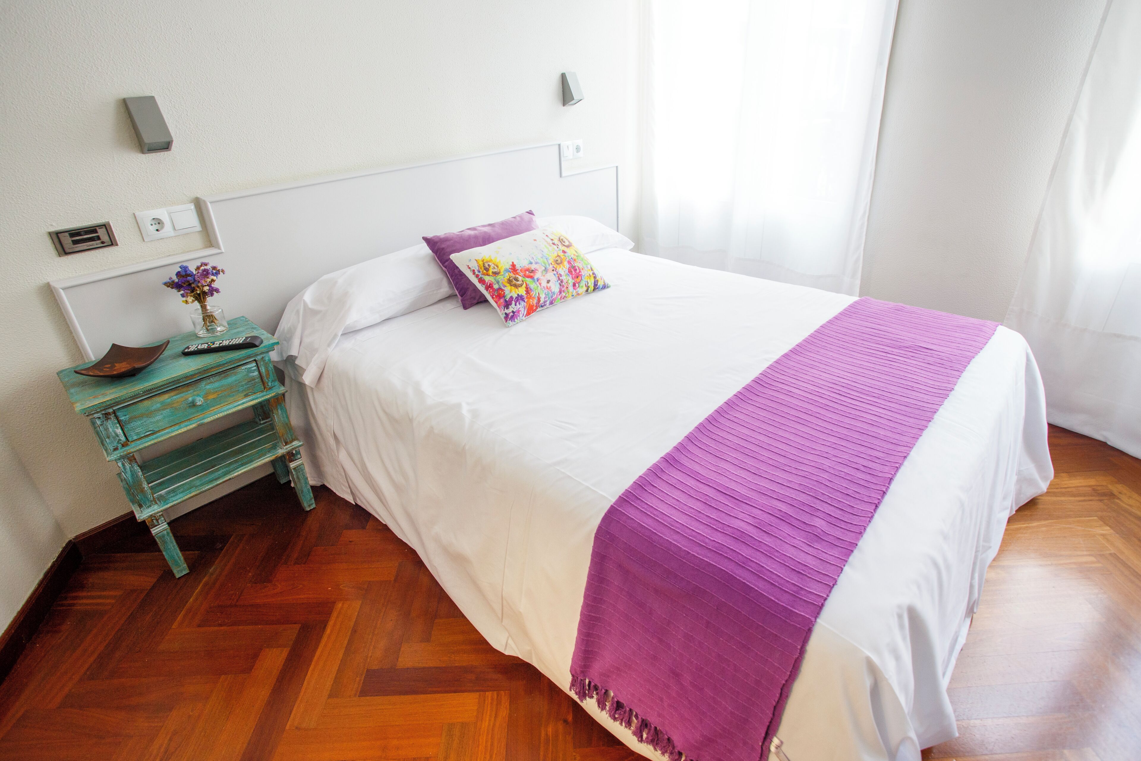 Un dormitorio con piso de madera, una cama con ropa blanca y una manta morada, una mesita de noche con un florero y un control remoto, y un aplique de pared.