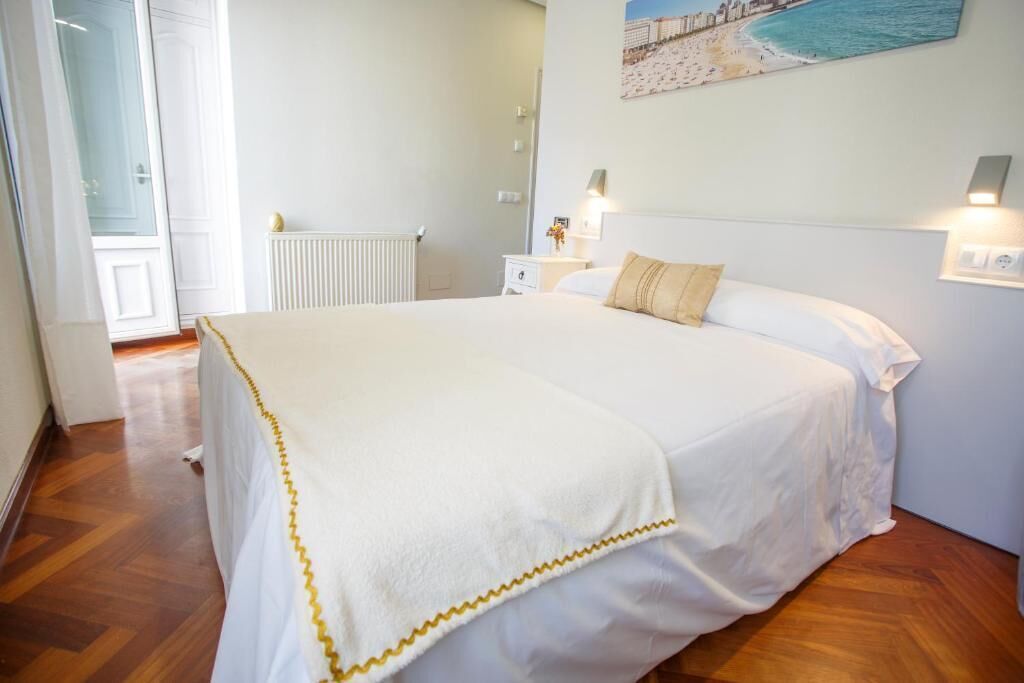 Una cama bien hecha con una colcha blanca y amarilla, una mesita de noche con una lámpara y un cuadro de una playa en la pared.