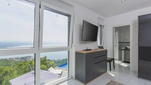 A modern hotel room with large windows, a flat-screen TV, a wooden desk, and a view of a city and water. - Villa Diamond in Opatija - Crovillas (Opatija)