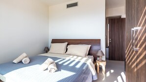 A neatly made bed with a blue quilt, two pillows, and rolled towels. A small bedside table with a lamp is beside the bed. - Elpida Luxury Villa, Private Pool, Fantastic View (Chania)